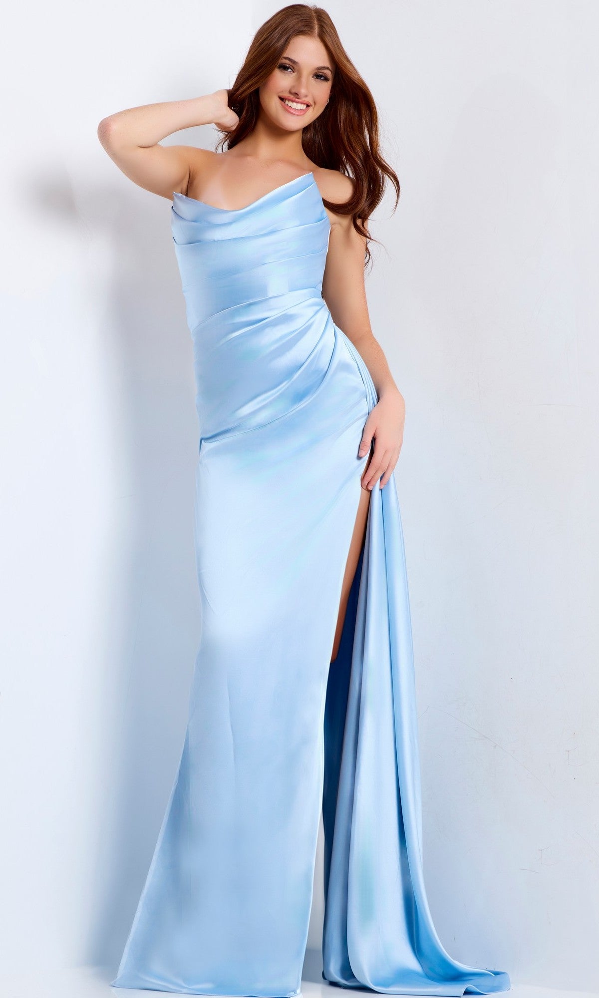 A woman in the Jovani 47160 long prom dress—light blue, strapless satin with a cowl neckline and high-leg side slit—stands against a plain background, smiling and posing with one hand behind her head.